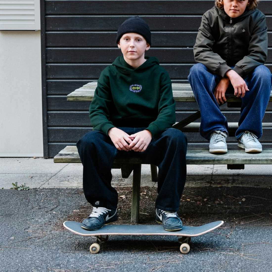 Enfant portant un sweat à capuche vert Born2ride Volcom Fa Ozzy Po Youth avec logo graphique, assis sur un banc